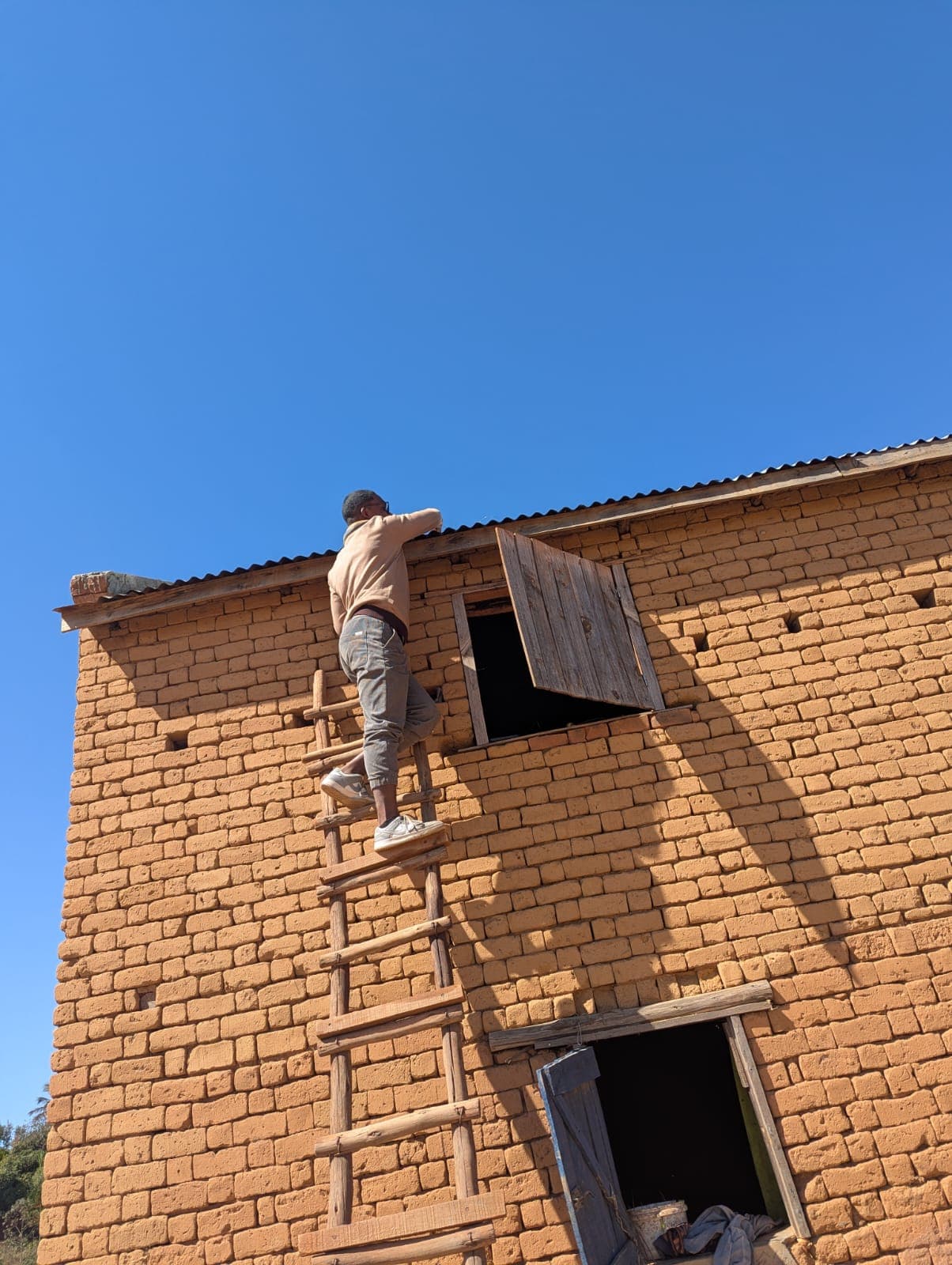 Un technicien Hazav'Iary monte à l'échelle vers la toiture d'une maison en briques de terre pour poser un panneau
