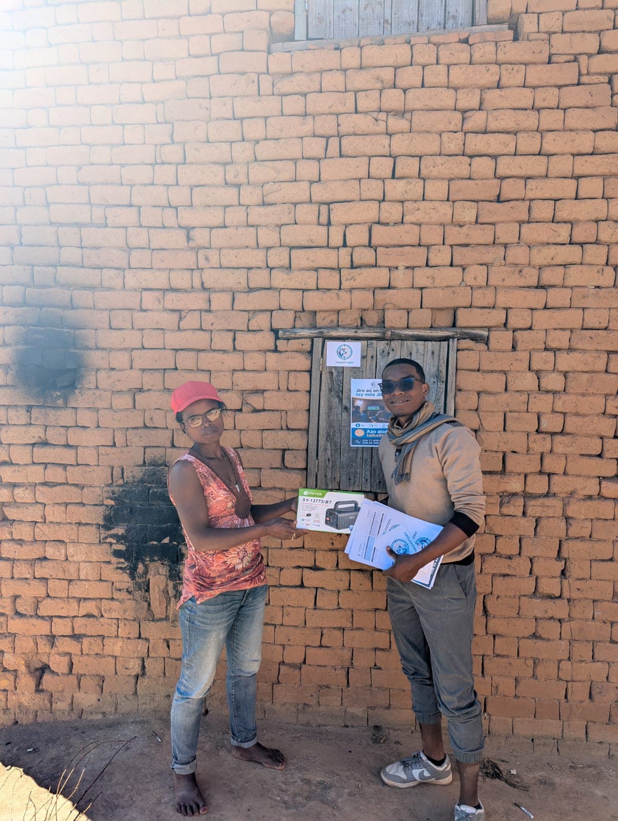 Remise d'un générateur solaire à une cliente devant sa maison, l'affiche Hazav'Iary posée sur le volet